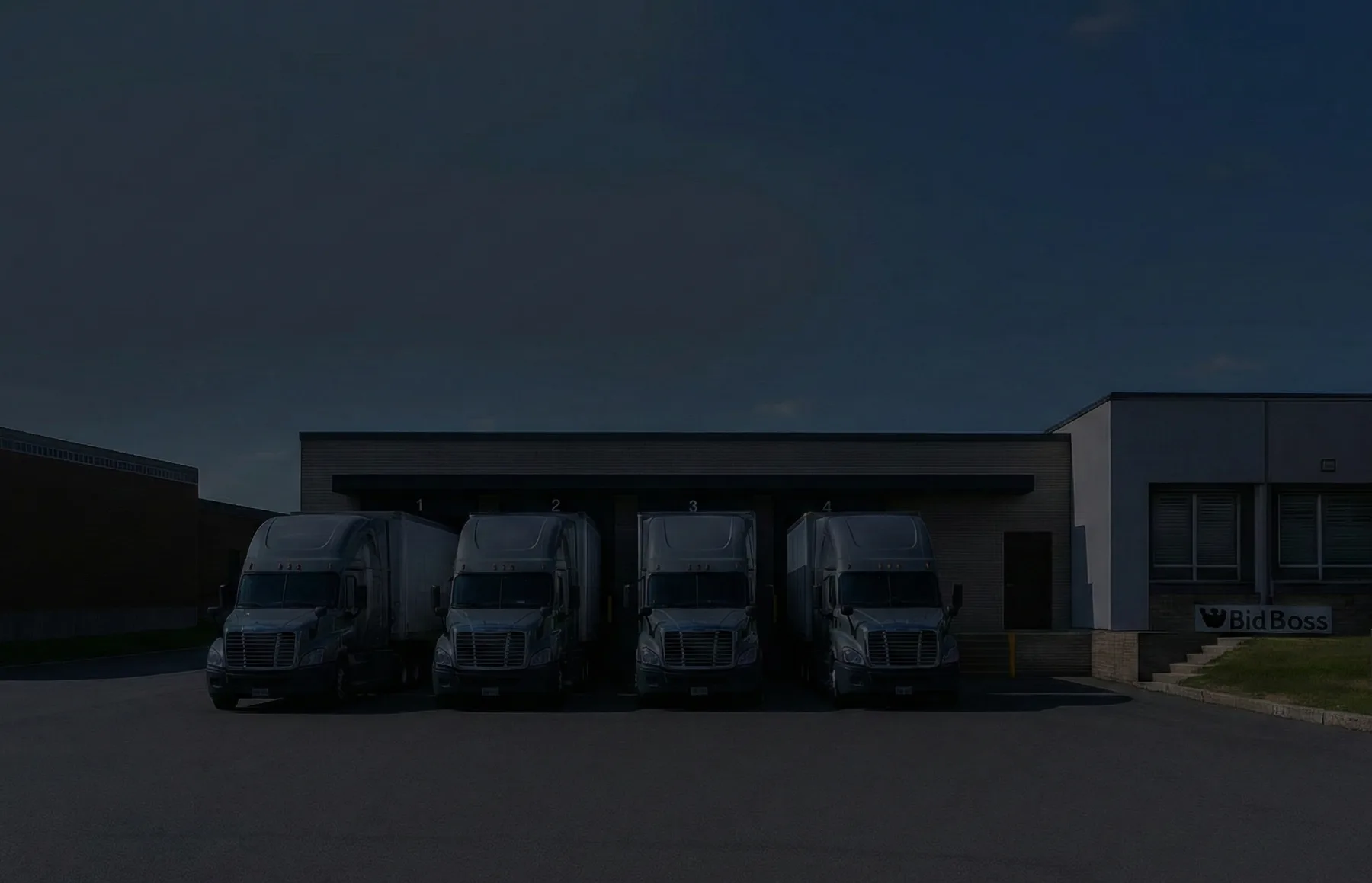 Four tractor units backed into numbered loading bays at dusk, with the Bid Boss sign on the building beside them.